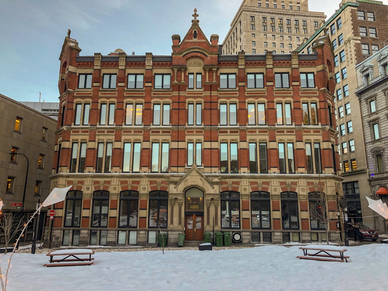 A red brick and sandstone building facing a snow-covered square with two picnic tables