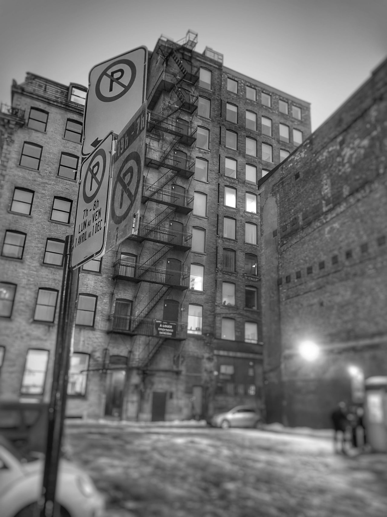 A black and white view of a brick building with a zigzag fire escape behind no-parking signs
