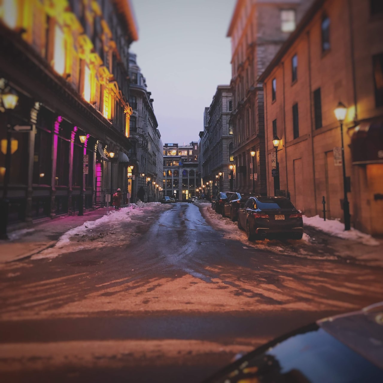 A narrow icy street between stone and brick buildings, lit by lanterns and street lamps