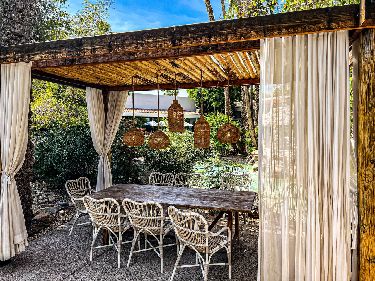 A timber cabana with sheer white curtains, woven pendant lamps and a long table with rattan chairs
