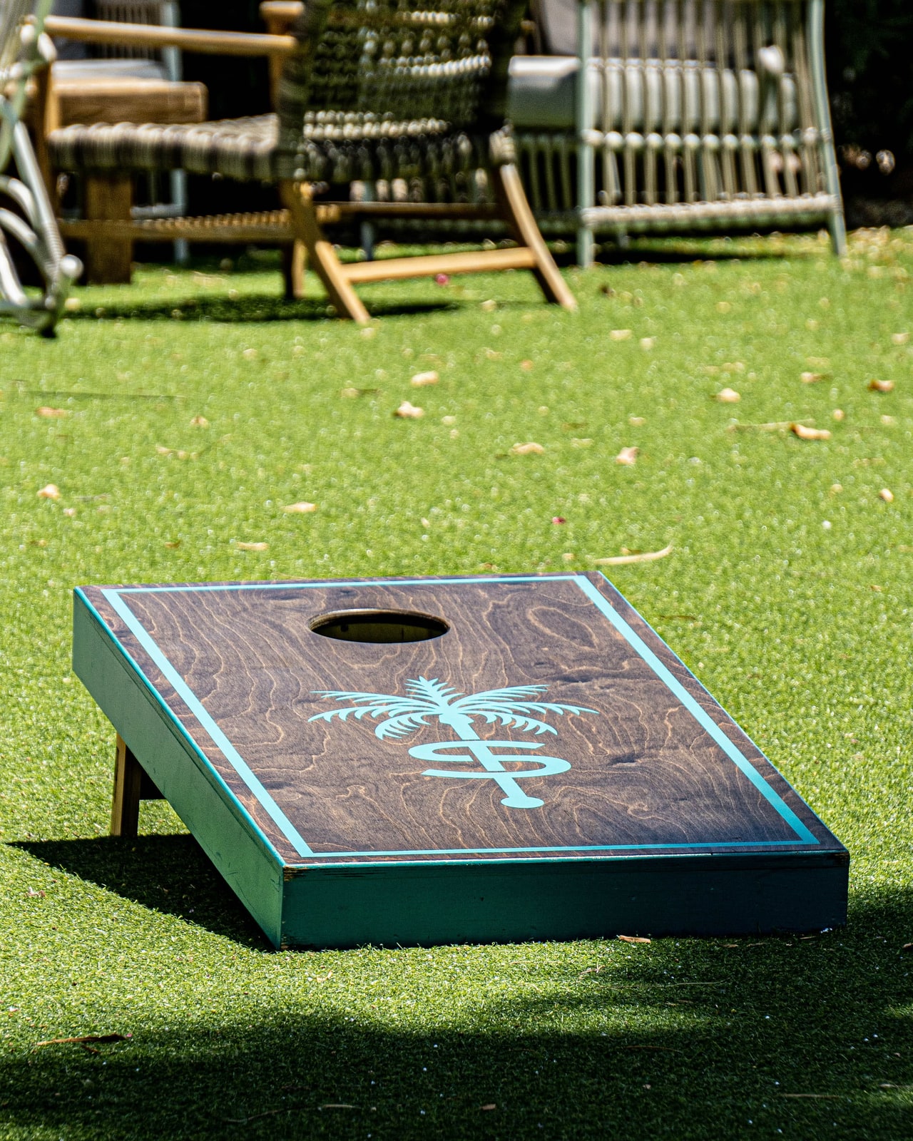 A dark wooden cornhole board with a turquoise border and palm-tree emblem on bright green turf