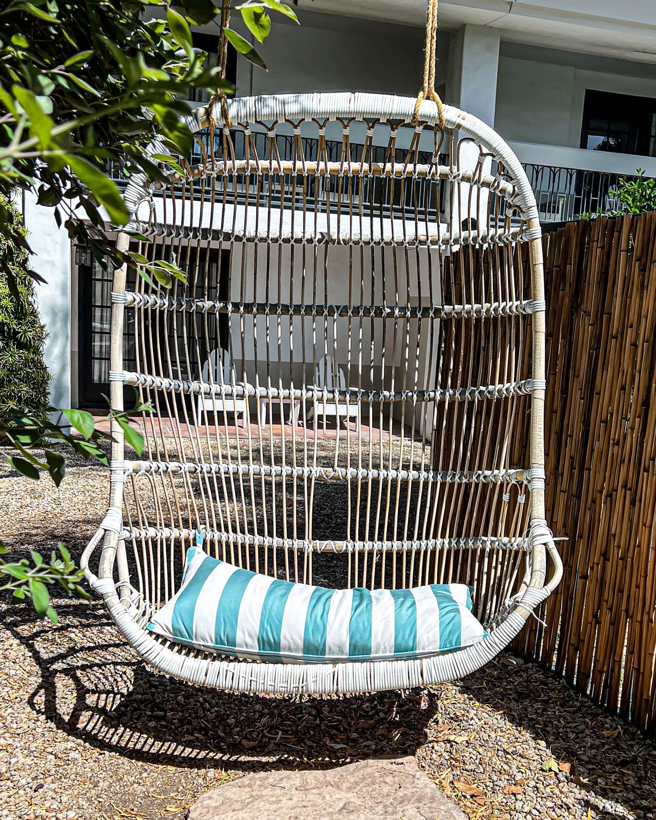 A white rattan hanging chair on two ropes, holding one turquoise and white striped cushion