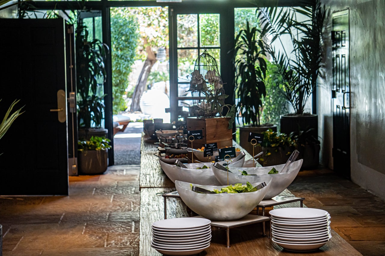 Pearly bowls of salad greens with tongs and stacked plates, glass doors open to a garden